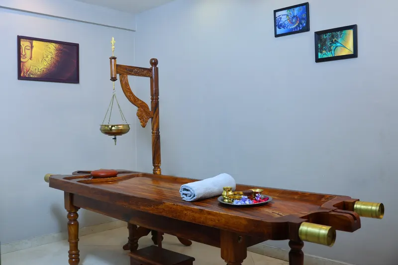 An Ayurveda treatment room with a carved wooden table, a folded towel and a brass oil vessel hanging above