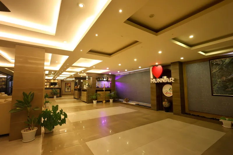 A wide lobby corridor with potted plants, a lit ceiling and an “I love Munnar” sign
